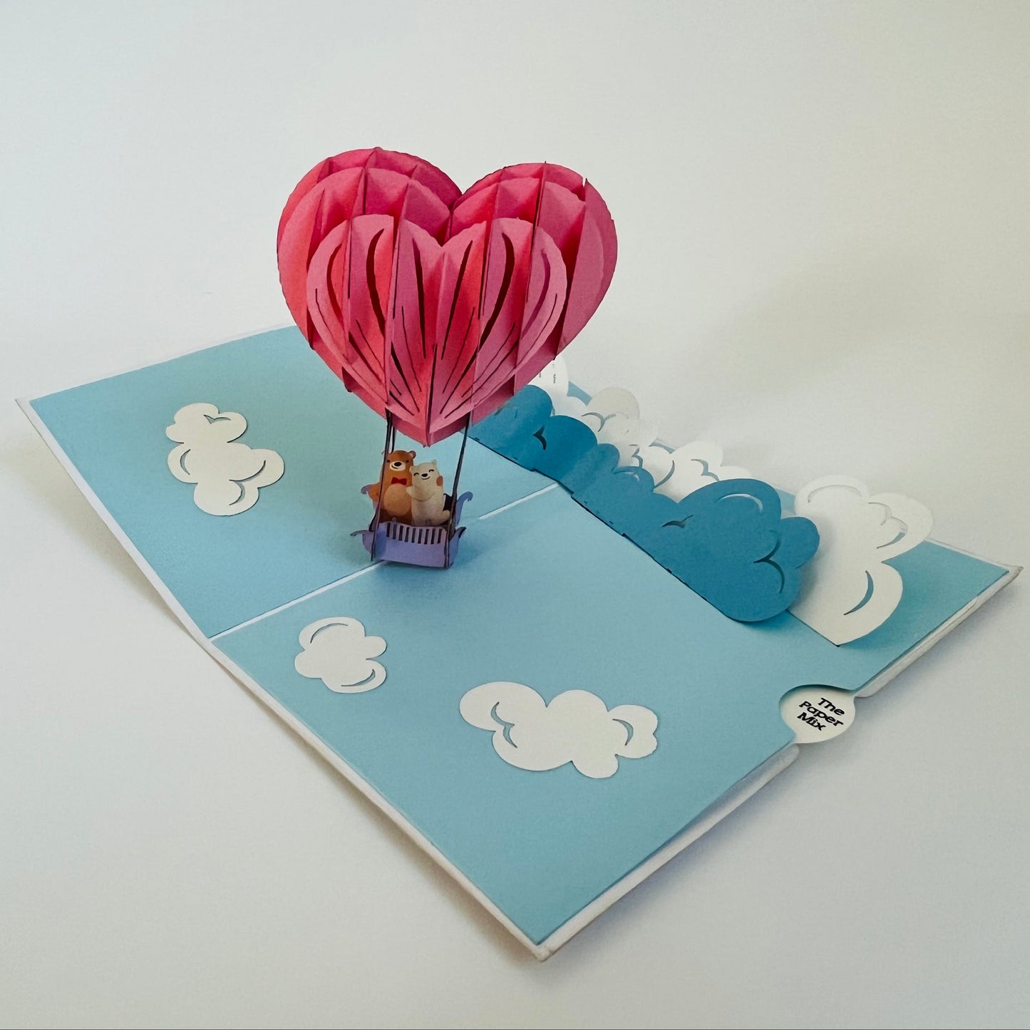 A handcrafted pop-up greeting card with a pink hot air balloon design, featuring a teddy bear inside the balloon, and blue sky with clouds.