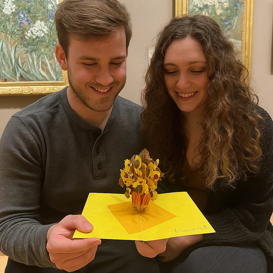 Two people holding a yellow pop-up card with a sunflower design in an art gallery setting.