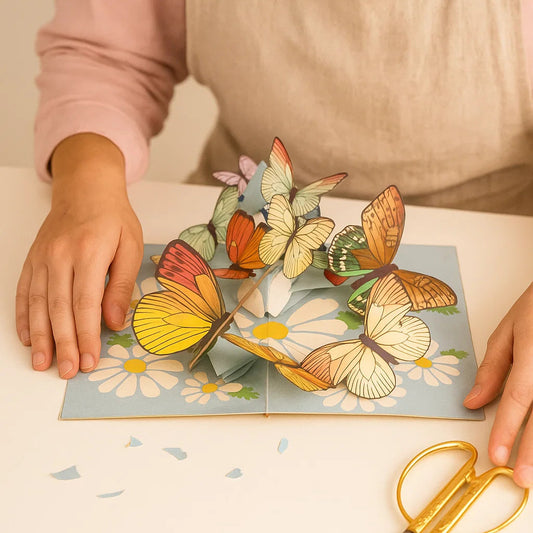 Person holding a pop-up card with butterflies and flowers, with a pair of scissors on a light surface.