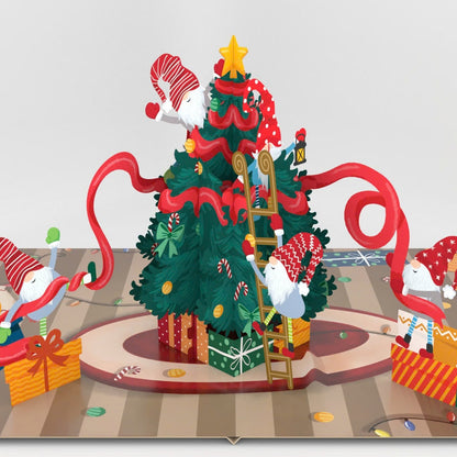 A 3D pop-up cardboard Christmas scene showing a decorated Christmas tree with Santa Claus, gifts, and festive decorations on a striped floor against a light gray background.