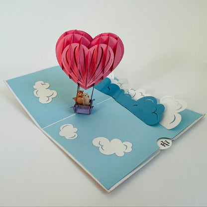 A handcrafted pop-up greeting card with a pink hot air balloon design, featuring a teddy bear inside the balloon, and blue sky with clouds.