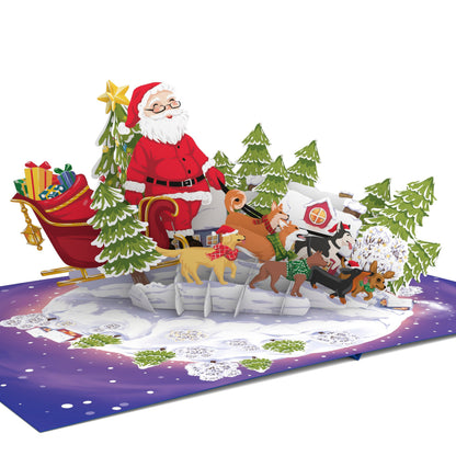 A 3D pop-up cardboard Christmas scene card featuring Santa Claus in his sleigh with reindeer, a decorated tree, and gifts on a snowy landscape base against a white background.