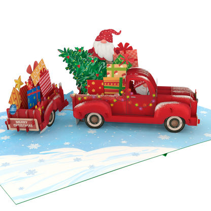 A 3D pop-up cardboard Christmas gift truck scene on a snowy blue ground. A red pickup truck carries a green Christmas tree and wrapped presents, with Santa Claus visible in the driver's seat.