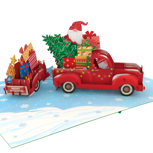 A 3D pop-up cardboard Christmas gift truck scene on a snowy blue ground. A red pickup truck carries a green Christmas tree and wrapped presents, with Santa Claus visible in the driver's seat.
