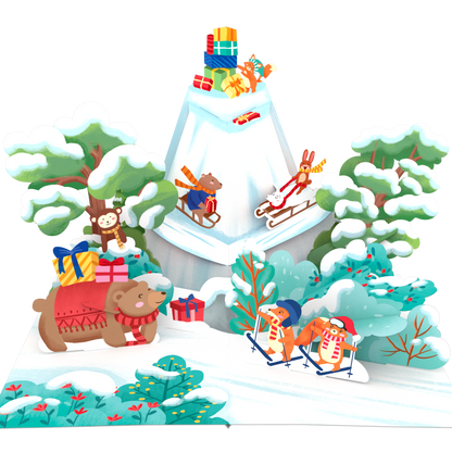 A 3D papercraft pop-up display featuring a winter Christmas scene with a snow-covered mountain, sleighs, a bear pulling a sled, a monkey riding a sled, and gift boxes, all set against a plain white background.