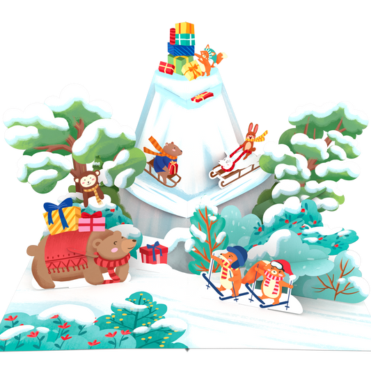 A 3D papercraft pop-up display featuring a winter Christmas scene with a snow-covered mountain, sleighs, a bear pulling a sled, a monkey riding a sled, and gift boxes, all set against a plain white background.