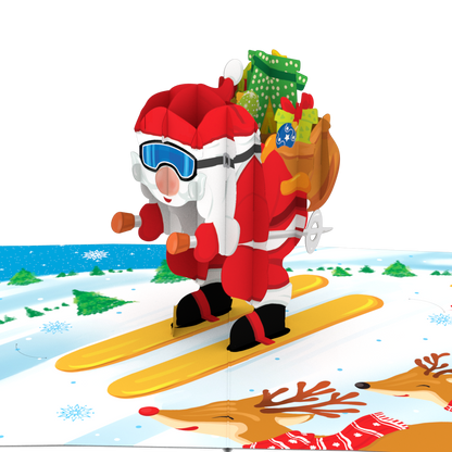 A 3D pop-up cardboard gift box featuring a festive Christmas winter scene. The design shows Santa Claus skiing down a snowy slope with a sack of gifts, accompanied by a penguin, reindeer, and pine tree, all set against a plain white background.