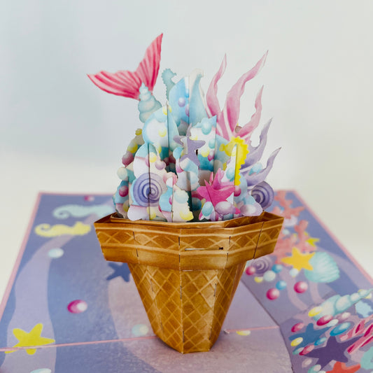 A handcrafted pop-up greeting card depicting a whimsical ice cream cone with mermaid tails and dreamy swirls in a purple unicorn hue on a patterned background.