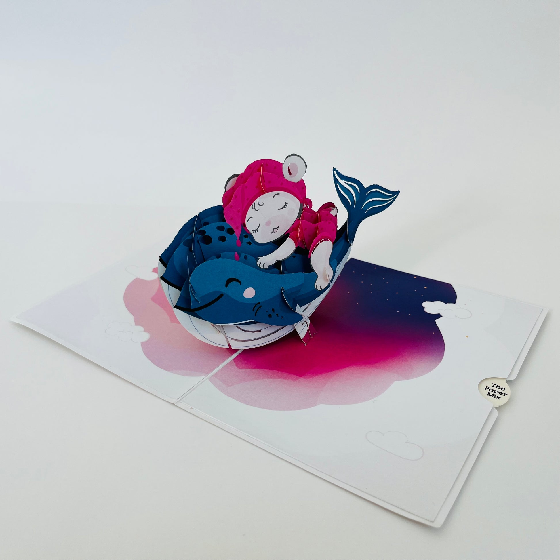 A pop-up greeting card depicting a blue whale with a person sleeping on it, set against a pink and white background.