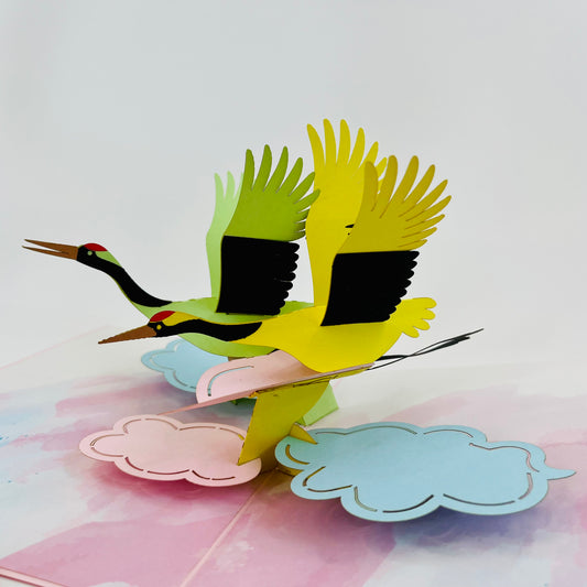 A colorful greeting card featuring a pop-up design with cranes flying over clouds.