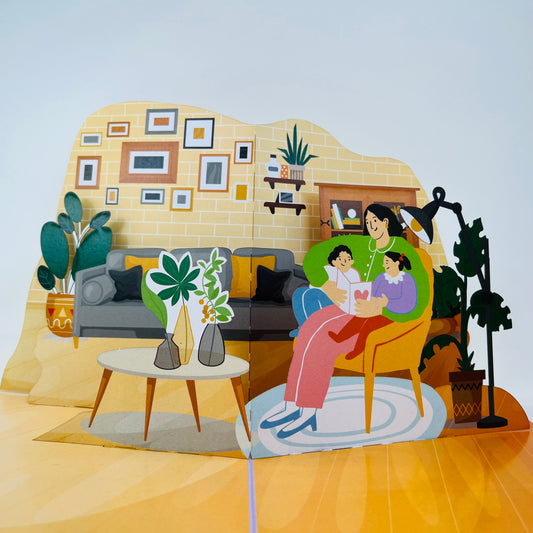 A 3D pop-up greeting card depicting a mother sitting with two children on a chair, reading a book. The card features detailed, colorful illustrations of a living room setting with decorations and plants.