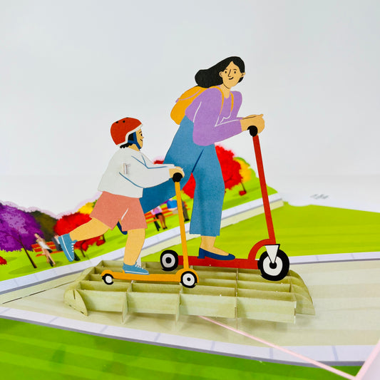A pop-up greeting card depicting a three-dimensional scene with a mother and child riding scooters. The card is colorful, featuring a green background, pink scooter, and red scooter with a detailed, playful design.