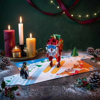 A festive tabletop scene with a small Santa Claus paper cutout wearing blue goggles and carrying gifts, standing on a printed snowy hill. A penguin figure is beside it, with lit candles, pinecones, ribbon, and holiday greenery around the setup.