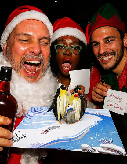 Three people in festive attire with a Christmas card featuring penguins.