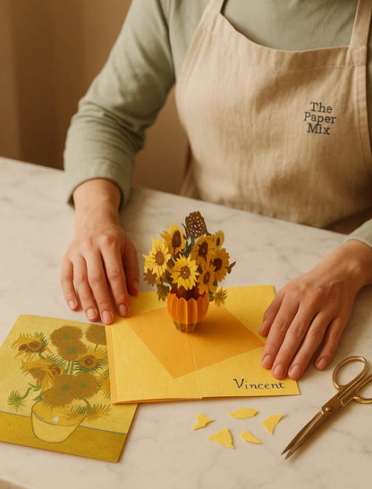 Person in an apron working with paper crafts on a table, including a 3D sunflower card and a small pumpkin-shaped sunflower display.